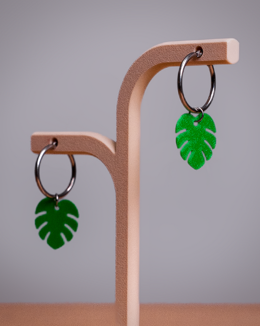 Green glitter monstera leaf drop earrings with silver metal rings, displayed on a light wood jewelry stand against a neutral gray background.