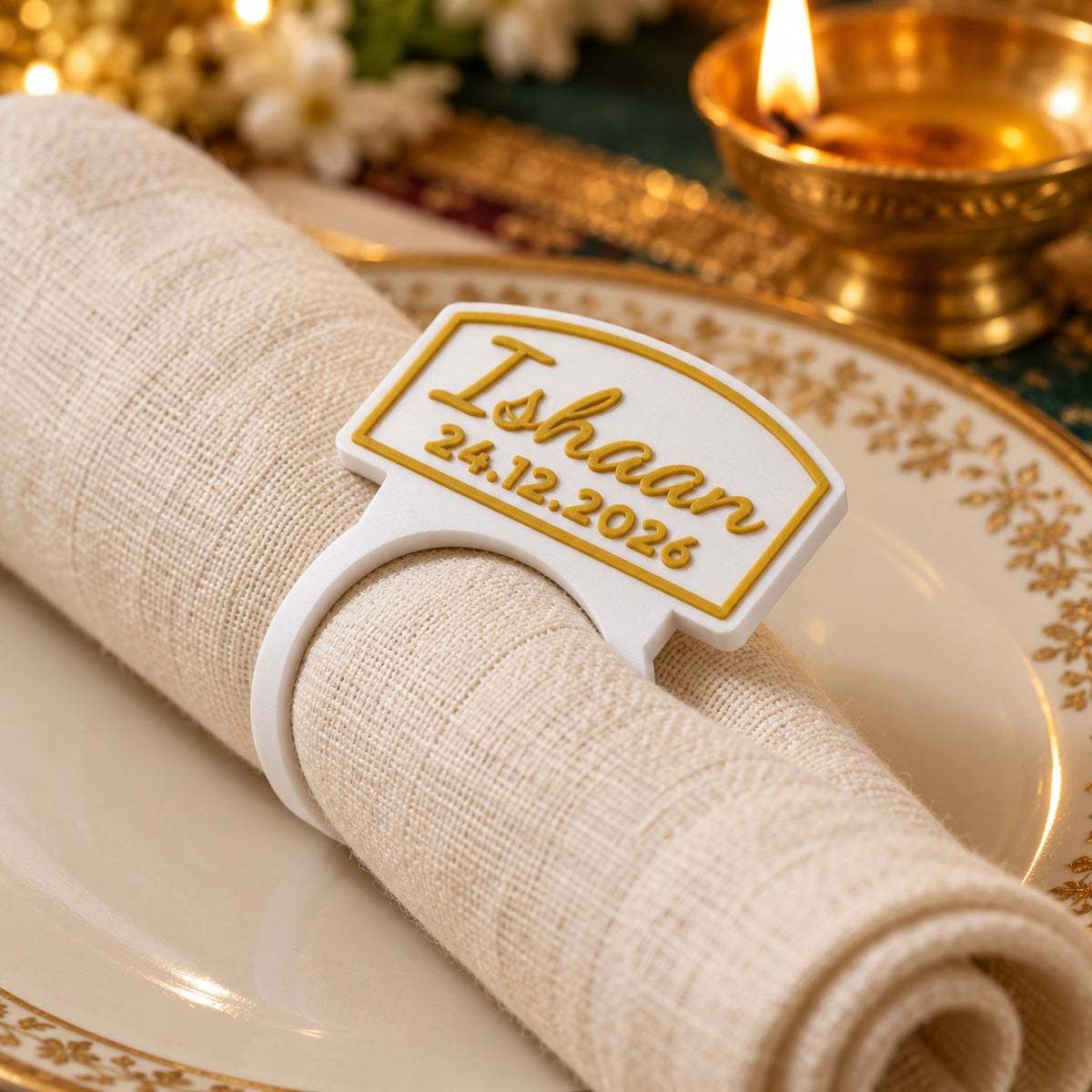 Personalised Napkin Ring: 'Ishaan' ring around a rolled ivory linen napkin on a dinner plate, on a festive dinner table with a brass diya softly out of focus