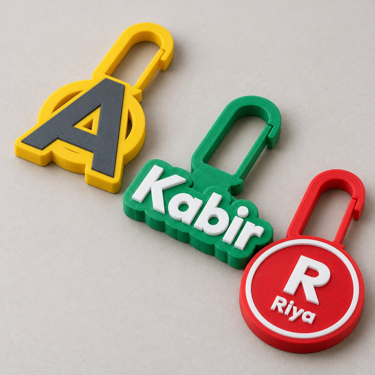 Three personalised zip charms: yellow initial A, green bubble name Kabir, red round badge R with Riya