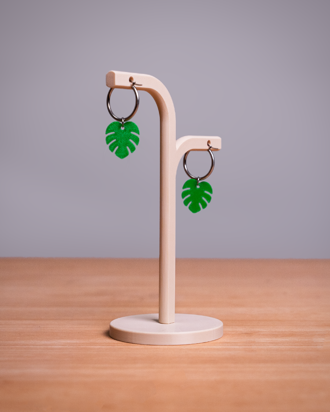 A pair of green monstera leaf drop earrings with silver-toned rings, displayed on a light wood T-stand against a neutral gray background and wooden tabletop.