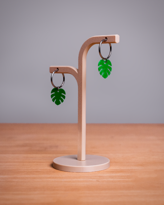 A pair of green monstera leaf hoop earrings displayed on a light wood stand on a wooden tabletop against a gray background.