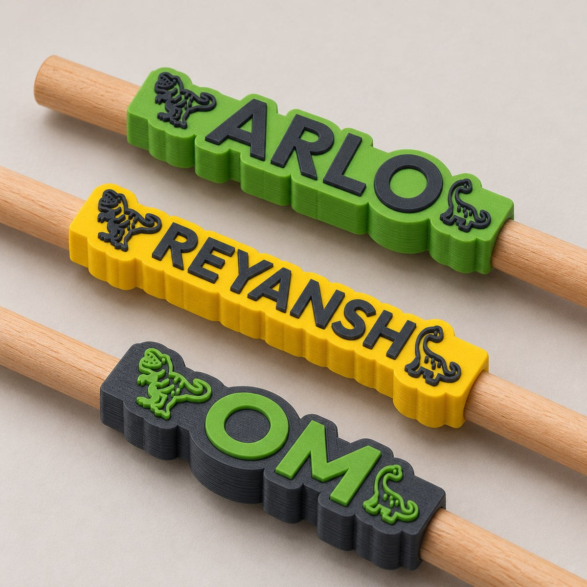 Dinosaur Name Pencil Topper: three toppers (Arlo, Reyansh, Om) each on the top of a pencil, the pencils lying side by side