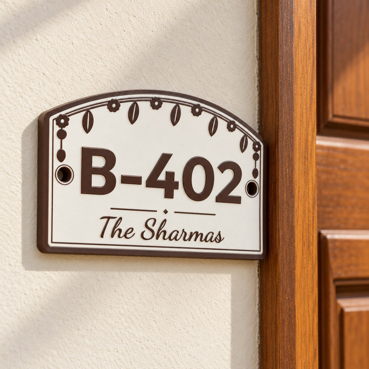 House Number Plate with Family Name: brown 'B-402 The Sharmas' plate mounted on a cream wall beside a wooden front door of an Indian apartment, soft daylight