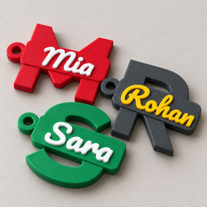 Initial and Name Banner Keychain: three keychains (M Mia, R Rohan, S Sara) laid flat, overlapping slightly, top-down at a slight angle