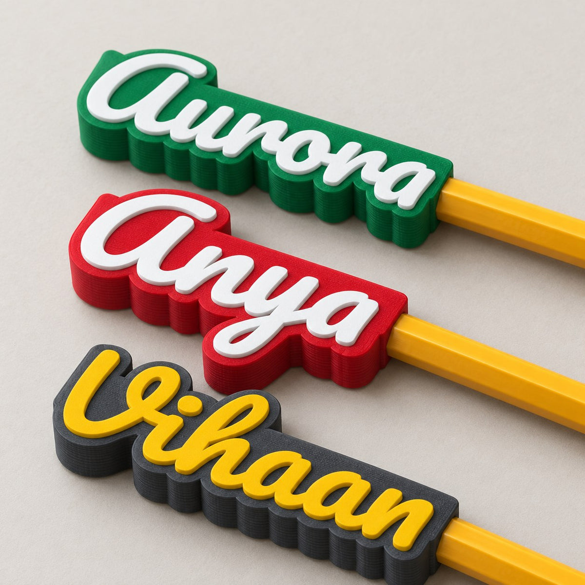 Name Pencil Topper: three toppers (Aurora, Anya, Vihaan) each pushed onto the top of a yellow HB pencil, the pencils lying side by side