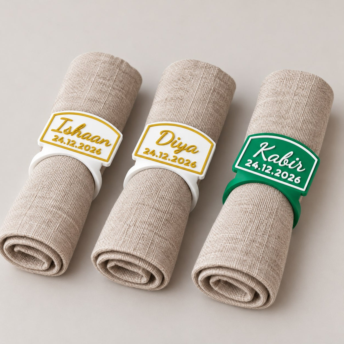 Personalised Napkin Ring: three napkin rings (Ishaan, Diya in white with gold letters, Kabir in green with white letters) each around a rolled linen napkin, lying in a row
