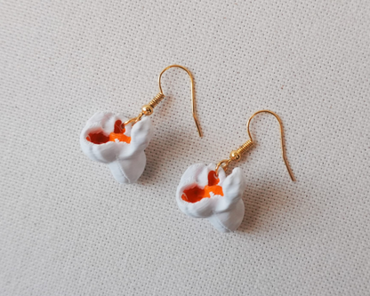 A pair of dangling earrings with white flower-shaped charms featuring orange centers, attached to gold fishhook wires, displayed on a textured off-white fabric background. The text 'Eyf_design' is visible in the bottom-left corner.