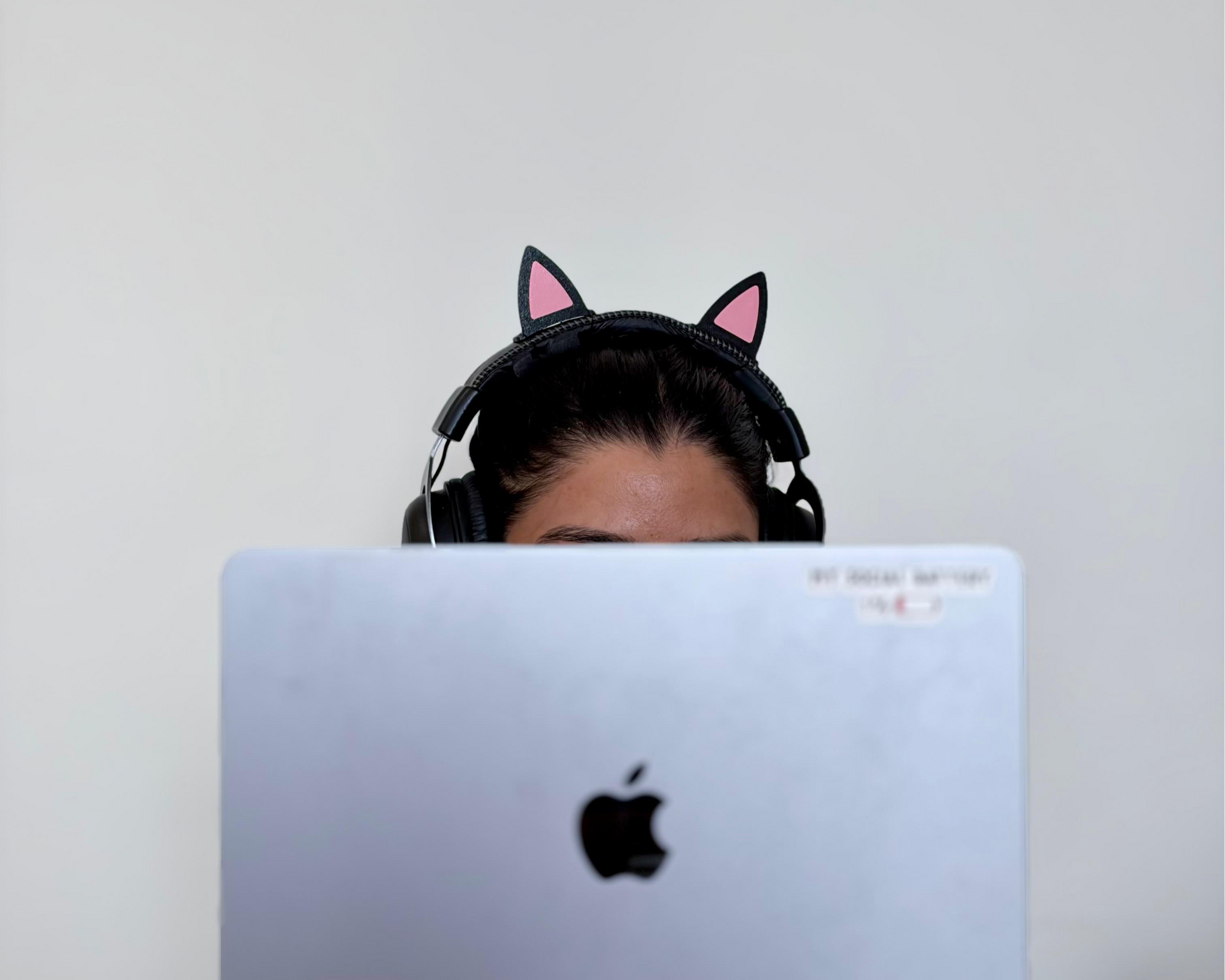 A person wearing black cat-ear headphones sits behind a silver laptop, with the Apple logo visible on the screen. The background is a plain light gray wall.