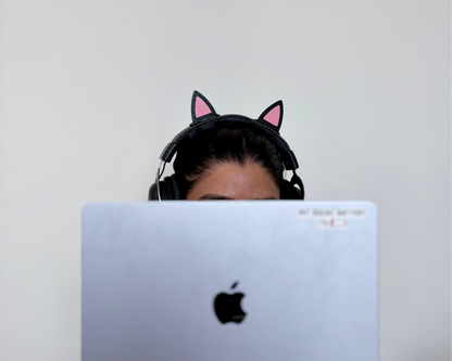 A person wearing black cat-ear headphones sits behind a silver laptop, with the Apple logo visible on the screen. The background is a plain light gray wall.