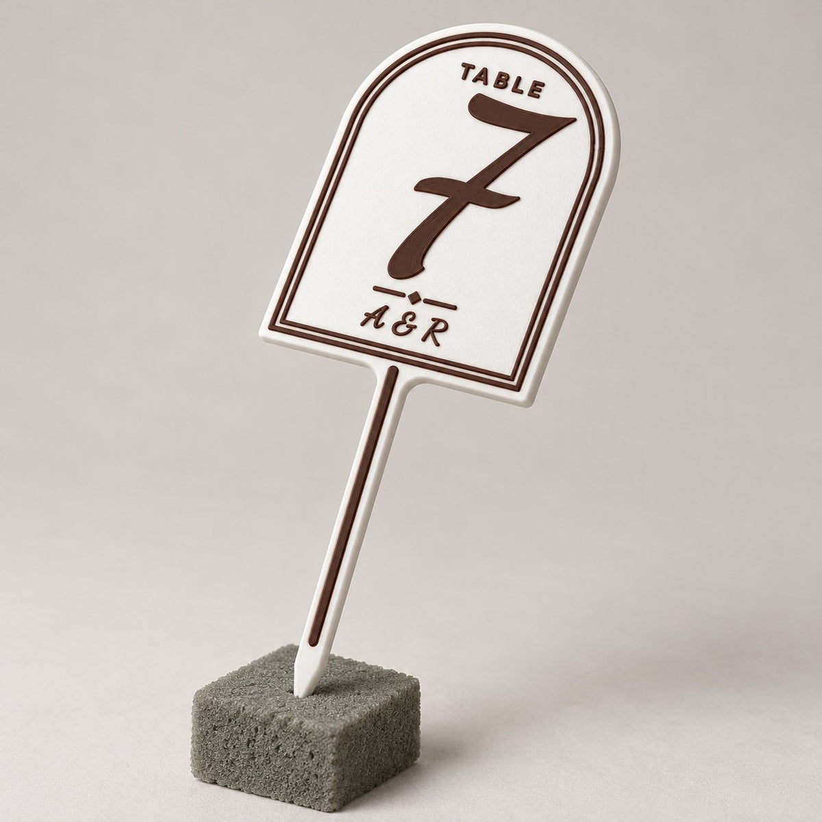 Wedding Table Number Stand: white table 7 stand standing upright pushed into a small block of grey florist foam, three-quarter angle, the whole stake visible