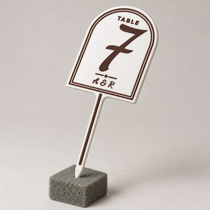Wedding Table Number Stand: white table 7 stand standing upright pushed into a small block of grey florist foam, three-quarter angle, the whole stake visible