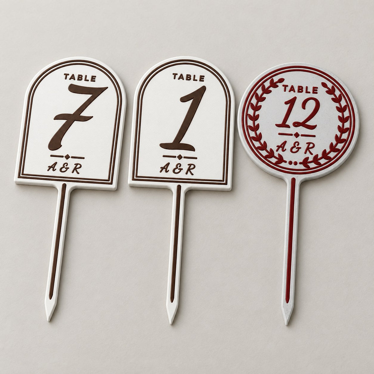 Wedding Table Number Stand: three stands (7 and 1 in white with brown letters, 12 in marble with deep red letters and a round wreath frame) lying flat side by side, top-down at a slight angle