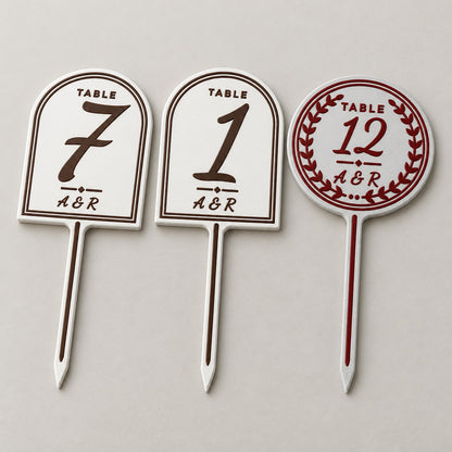 Wedding Table Number Stand: three stands (7 and 1 in white with brown letters, 12 in marble with deep red letters and a round wreath frame) lying flat side by side, top-down at a slight angle