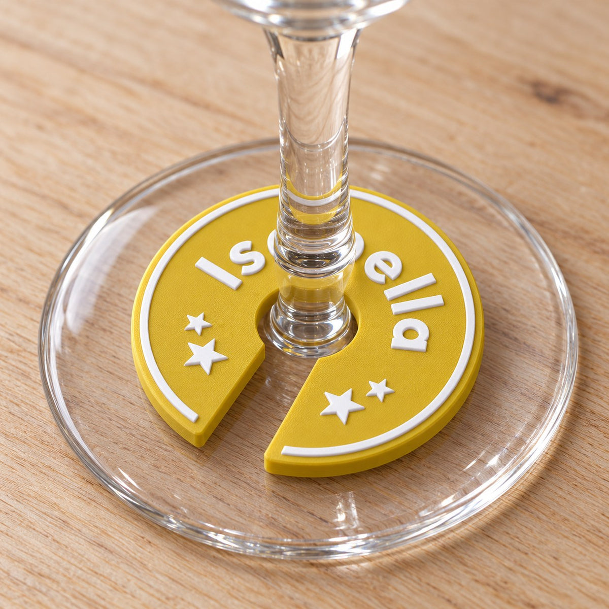 Wine Glass Name Tag: gold 'Isabella' ring clipped round the stem of a clear wine glass, resting flat on the glass foot, on a light wooden table