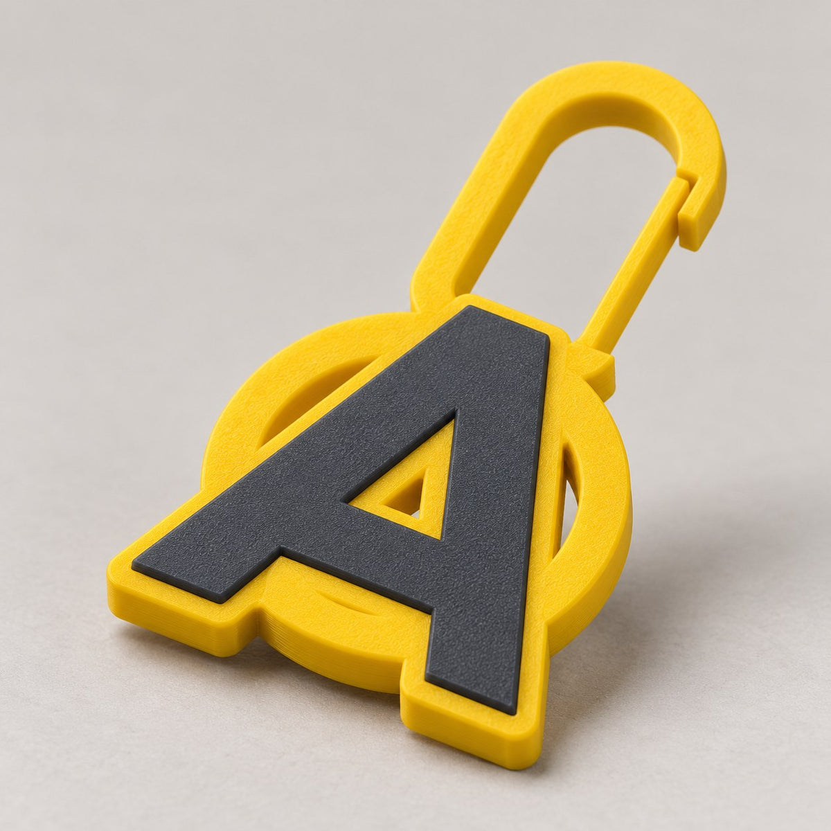Yellow 3D-printed zip charm with a dark grey letter A in a ring and a clip hook
